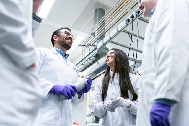 Chemical engineers wearing white labcoats, bright purple gloves, and safety glasses, talk in laboratory. Younger woman with long dark hair is looking up at a bearded man holding a gas canister.