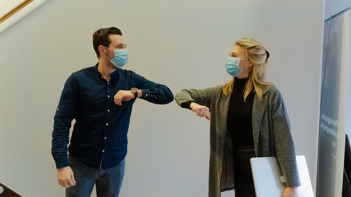 A man and a woman in business casual attire and wearing blue surgical masks are bumping elbows while social distancing. They are smiling from behind their masks as they greet each other.