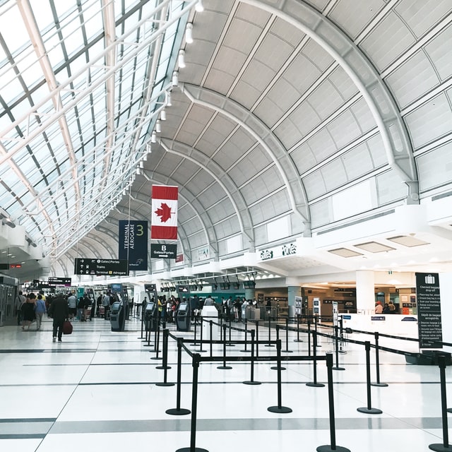 Image of Toronto Pearson Airport, indoors, a Canadian flag hangs above a nondescript group of travellers in the distance engaged in the hustle and bustle of air travel.