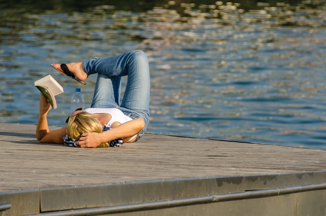 woman laying relaxed on her back, reading a book on a dock by the lake in summer