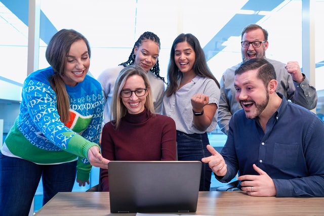 Group of diverse young people with excited facial expressions surround a laptop, some pump their fists, others point at the screen.